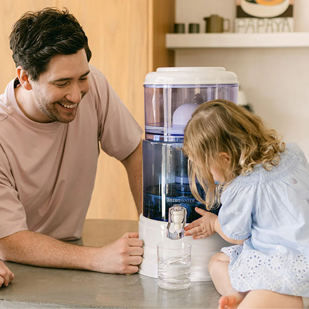 A smiling man watches a young girl press the tap on a zazen Alkaline Water System to fill a glass of water at the kitchen bench.